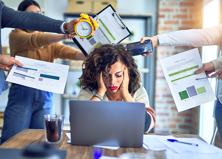 Stressed woman with face in hands at computer while others hand her deadlines