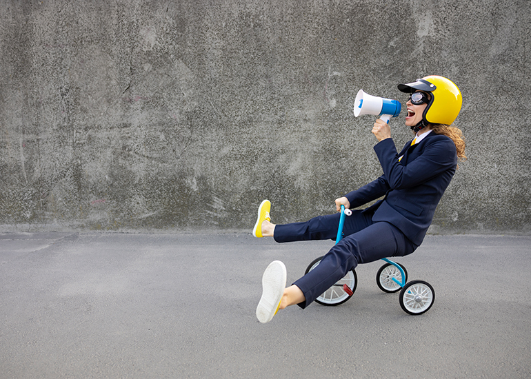 Happy business woman riding a childโs tricycle wearing a yellow helmet