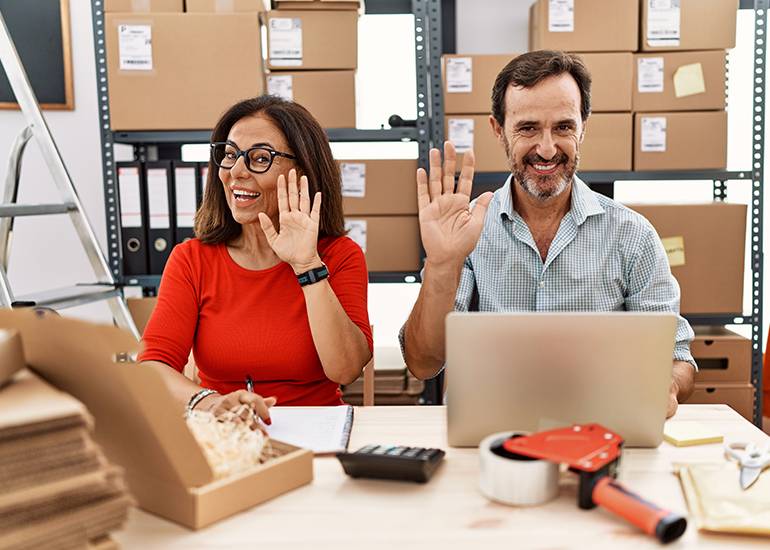 Business owner couple waving and smiling as they work on accounts and orders