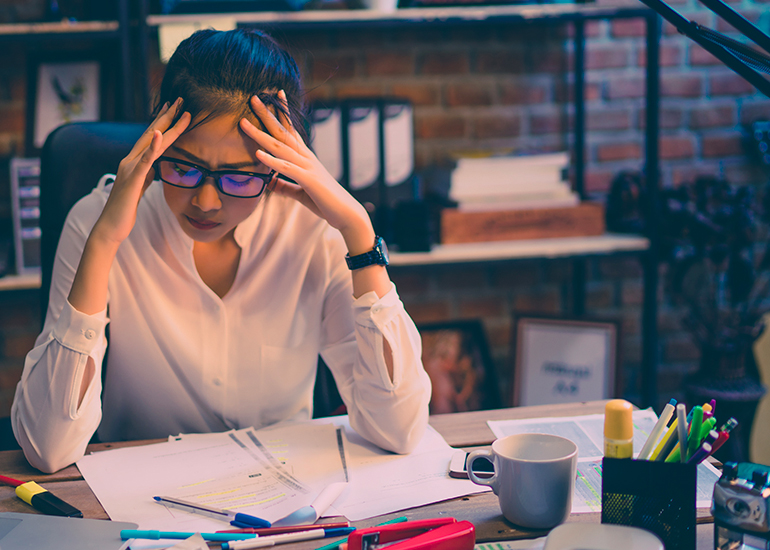 Stressed woman at desk with head in hands and paperwork strewn everywhere