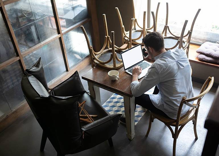 Man sitting in empty bar or cafe with laptop on tables and upturned chairs nearby