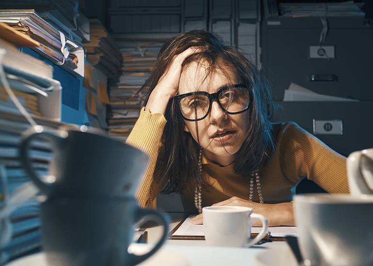 Busy, stressed woman sitting at desk surrounded by piles of paperwork