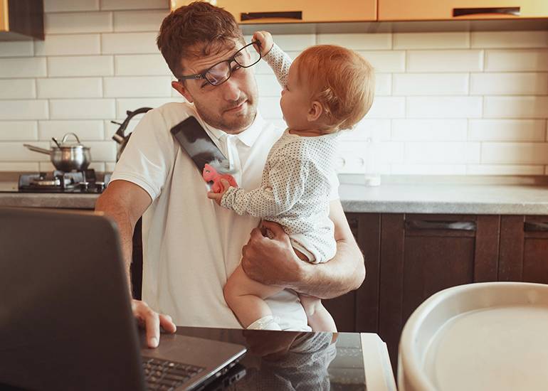 Dad working at laptop at home while baby pulls off his glasses