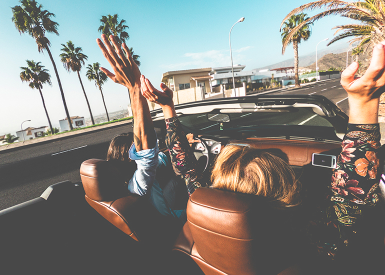 Two women driving in convertible car with hands in air