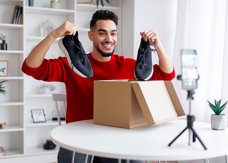 Young man holding shoes above box as he smiles into phone camera