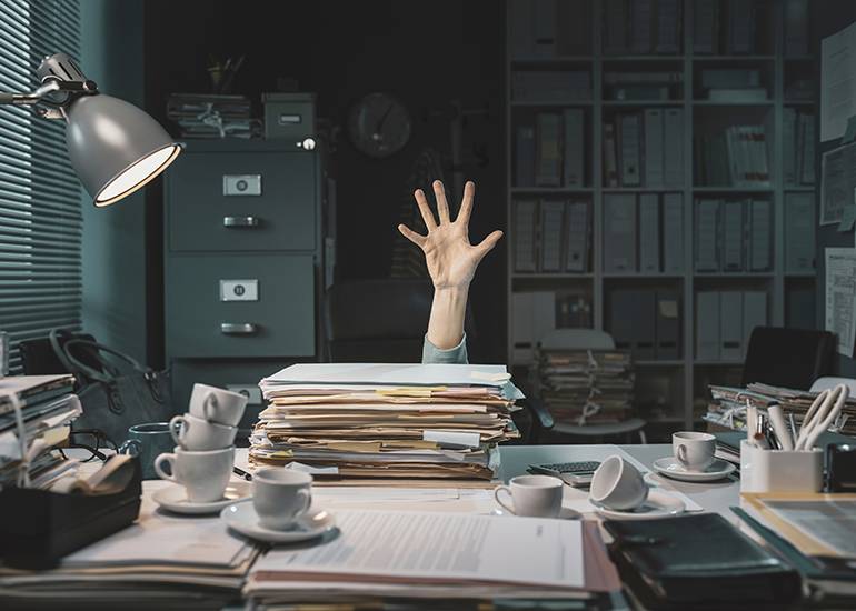 Overflowing desk with hand reaching up for help from behind