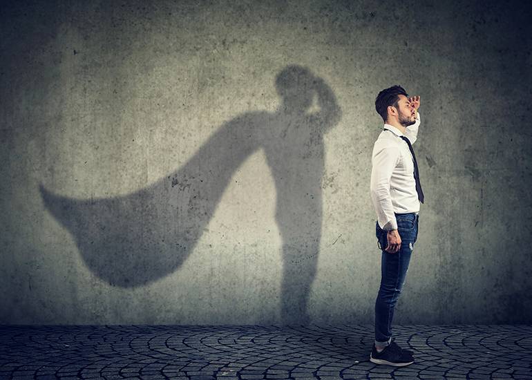 An ordinary looking business man in whit shrits and dark trousers stands side on - he looks to the horizon - bhdin him his shadow shows a siperhero with a cape unfurling. Socia media branding concept