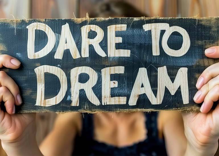 a woman holds up a sign that reads dare to dream in front of her face: bold concept.