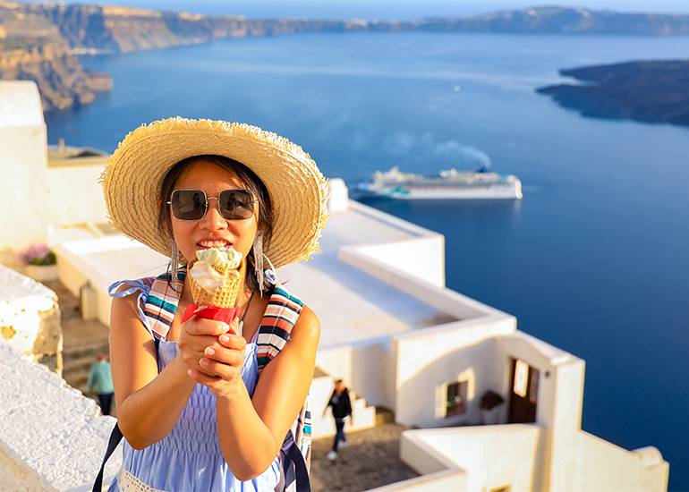 A woman eats an icecream contemplating a business idea. In the background we see blue wtaer and white stone buildings in a greecian seaside town