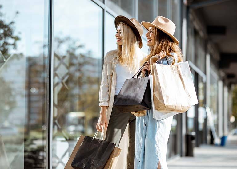 Two happy girlfriends looking in the shop window while standing with shopping bags near the mall